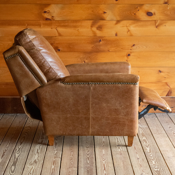 The reclined profile view of a muted brown leather recliner with nailhead trim against a wooden wall