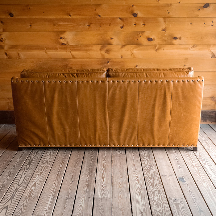 Back view of a full grain leather two-cushion sofa with rolled arms and nailhead trim on a wooden floor against a wooden wall