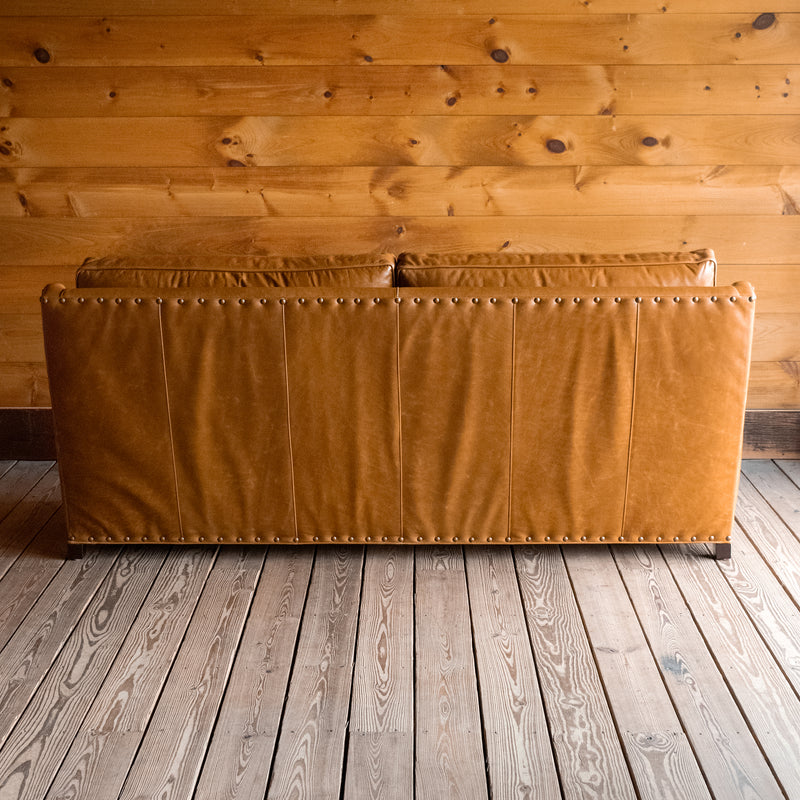 Back view of a full grain leather two-cushion sofa with rolled arms and nailhead trim on a wooden floor against a wooden wall