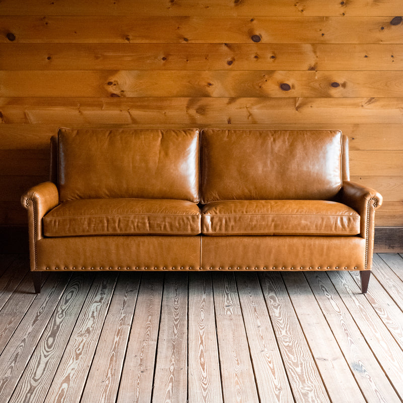 Front view of a full grain leather two-cushion sofa with rolled arms and nailhead trim on a wooden floor against a wooden wall