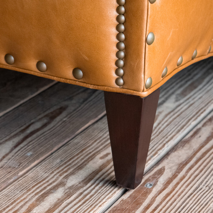 Leg close-up of a full grain leather two-cushion sofa with rolled arms and nailhead trim on a wooden floor