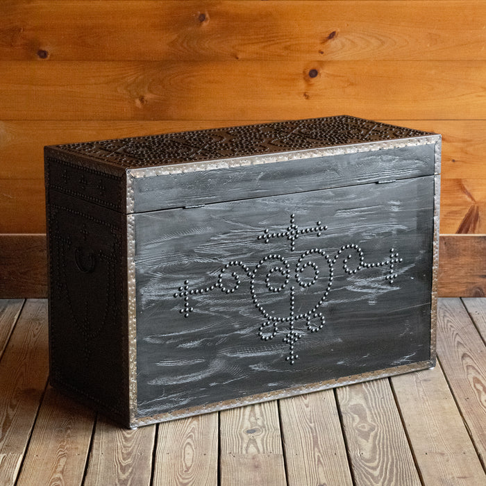 Angled back view of a dark reclaimed pine storage trunk, decorated with nailheads, on a wide-plank wooden floor against a rustic wooden wall 