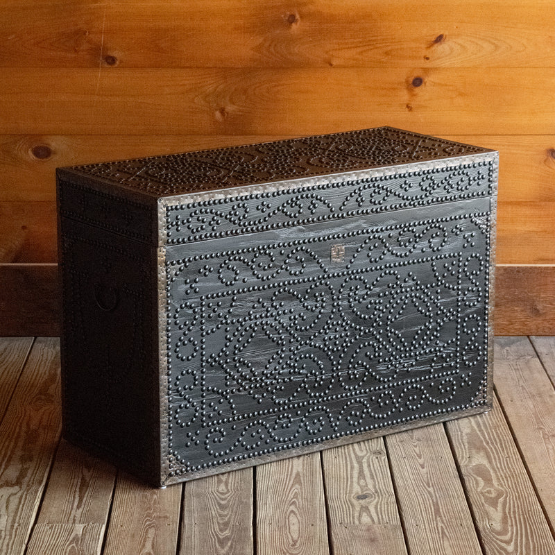 Angled front view of a dark reclaimed pine storage trunk, decorated with nailheads, on a wide-plank wooden floor against a rustic wooden wall 