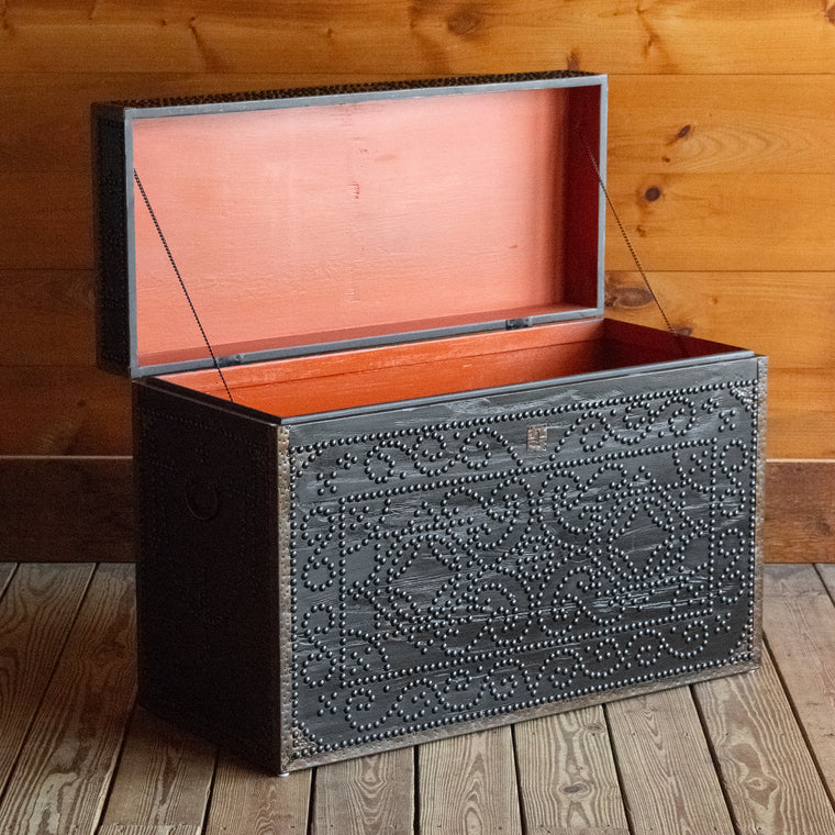 Angled back view of a dark reclaimed pine storage trunk, decorated with nailheads, on a wide-plank wooden floor against a rustic wooden wall 