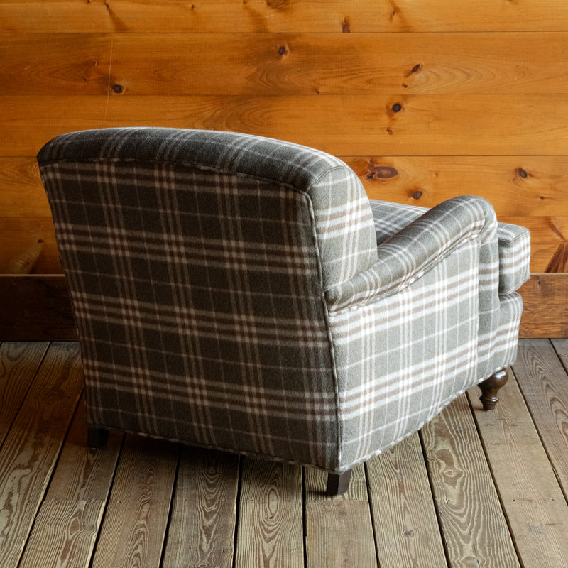 Angled back view of an oversized English rolled arm chair upholstered in a plaid wool fabric on a wide-plank wooden floor against a rustic wooden wall