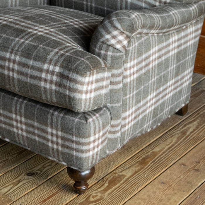 Close-up of the arm, cushion, and leg of an oversized English rolled arm chair upholstered in a plaid wool fabric on a wide-plank wooden floor