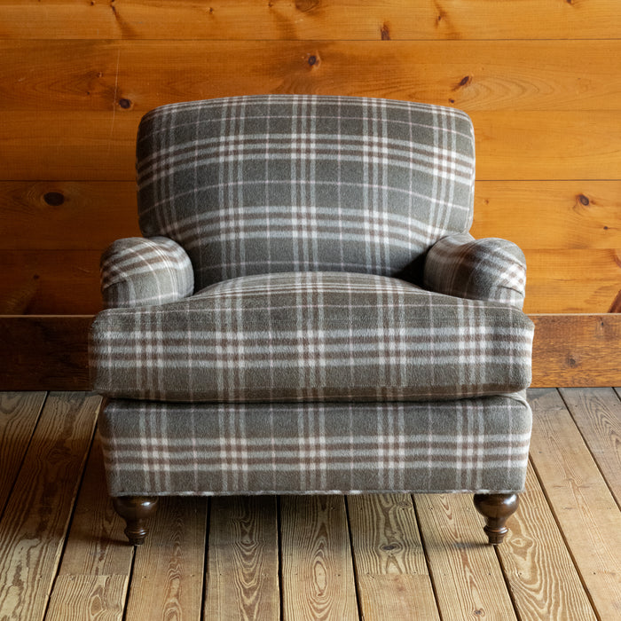 Front view of an oversized English rolled arm chair upholstered in a plaid wool fabric on a wide-plank wooden floor against a rustic wooden wall
