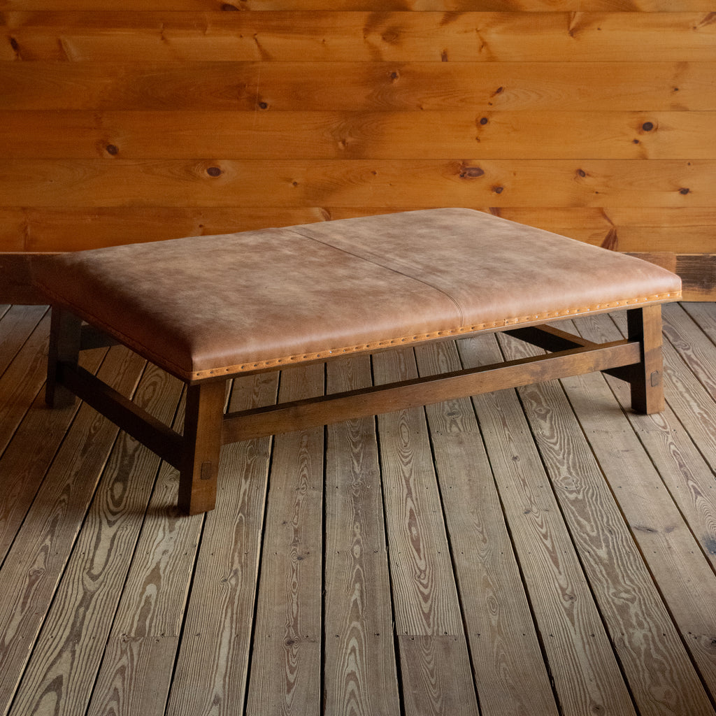 Angled front view of a rustic leather ottoman with an open hardwood frame on a wide-plank wooden floor against a rustic wooden wall
