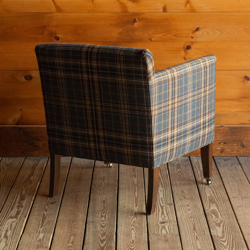 Angled back view of a low, tight back chair with track arms upholstered in a rustic plaid fabric on a wide-plank wood floor against a rustic wooden wall