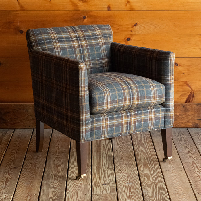 Angled front view of a low, tight back chair with track arms upholstered in a rustic plaid fabric on a wide-plank wood floor against a rustic wooden wall
