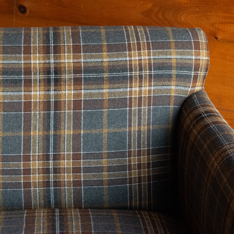 Angled back view of a low, tight back chair with track arms upholstered in a rustic plaid fabric on a wide-plank wood floor against a rustic wooden wall