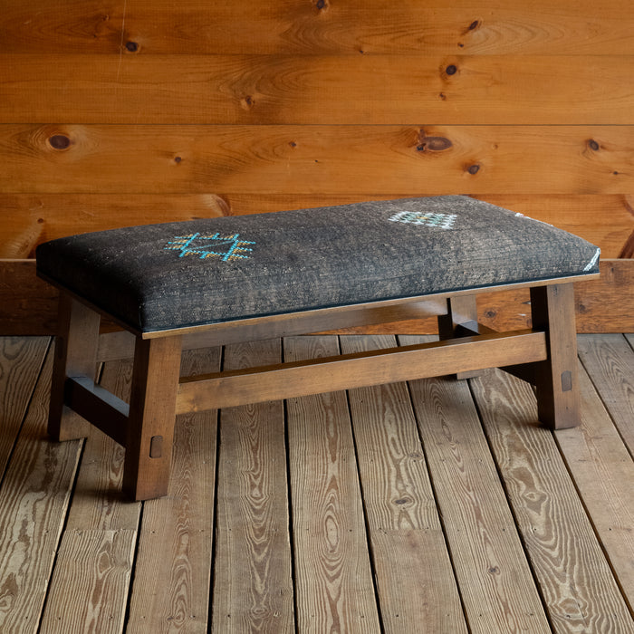 Angled front view of an ottoman with an open wooden frame upholstered in Moroccan cactus silk on a wide-plank wooden floor against a rustic wooden wall