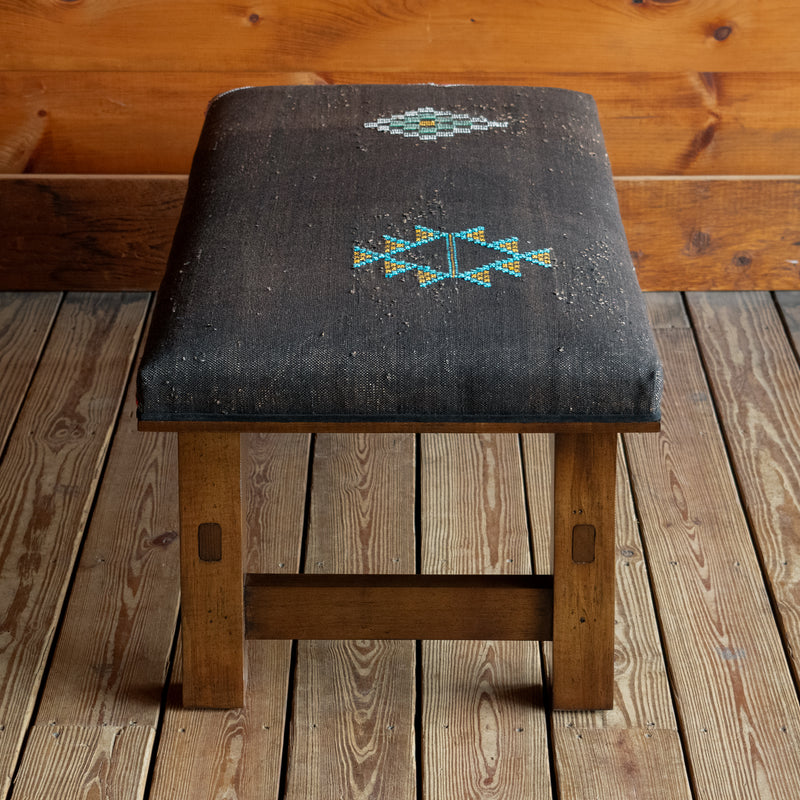 Profile view of an ottoman with an open wooden frame upholstered in Moroccan cactus silk on a wide-plank wooden floor against a rustic wooden wall