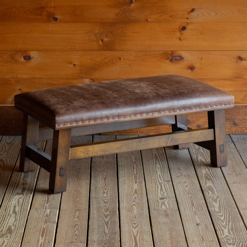 Angled front view of an ottoman upholstered in rustic leather with an open hardwood frame on a wide-plank wooden floor against a wooden wall