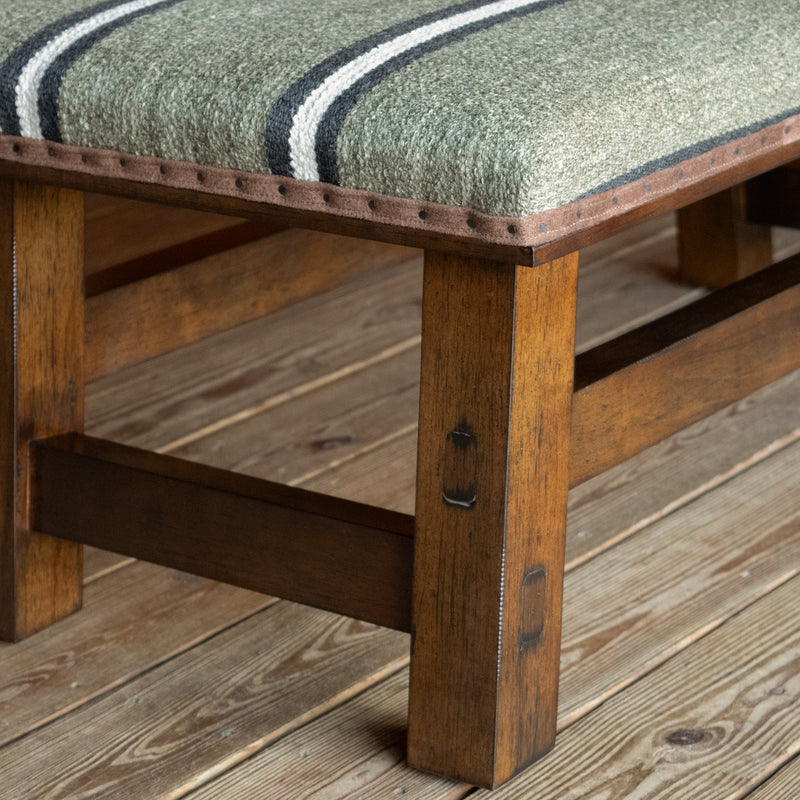 Close-up of the wooden base of a rustic ottoman upholstered in an olive stripe fabric on a wide-plank wooden floor