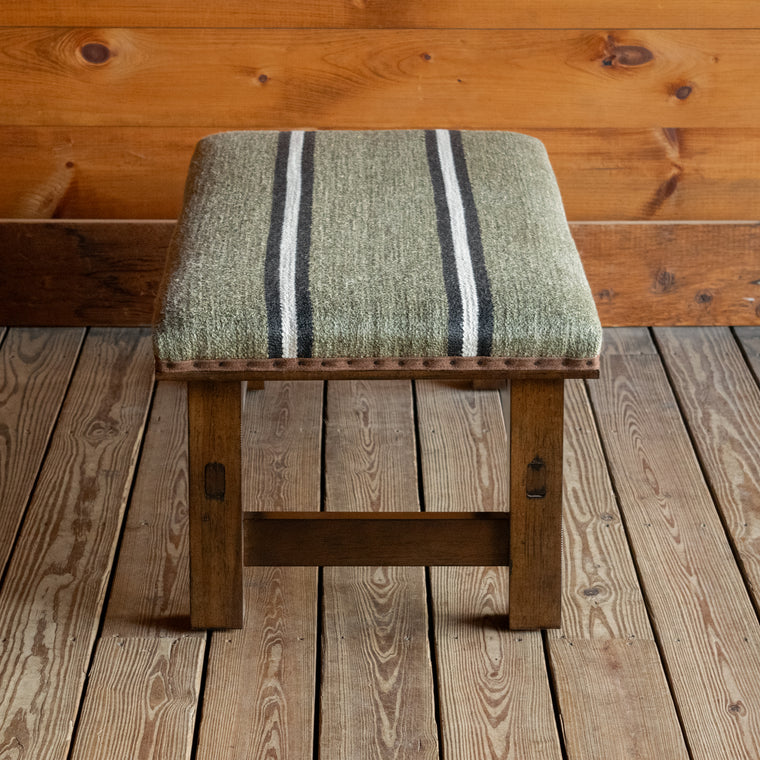 Close-up of the wooden base of a rustic ottoman upholstered in an olive stripe fabric on a wide-plank wooden floor