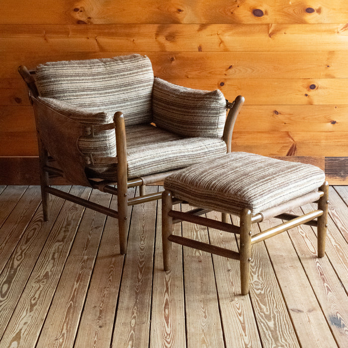 Angled front view of a safari-inspired chair and ottoman with leather sides and striped cushions on a wooden floor against a wooden wall
