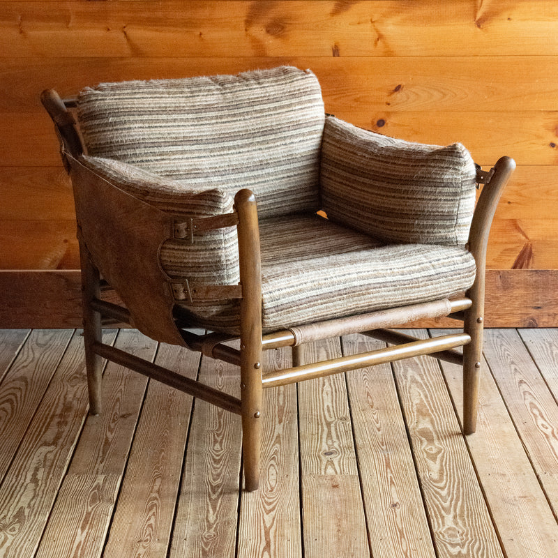 Angled front view of a safari-inspired chair with leather sides and striped cushions on a wooden floor against a wooden wall