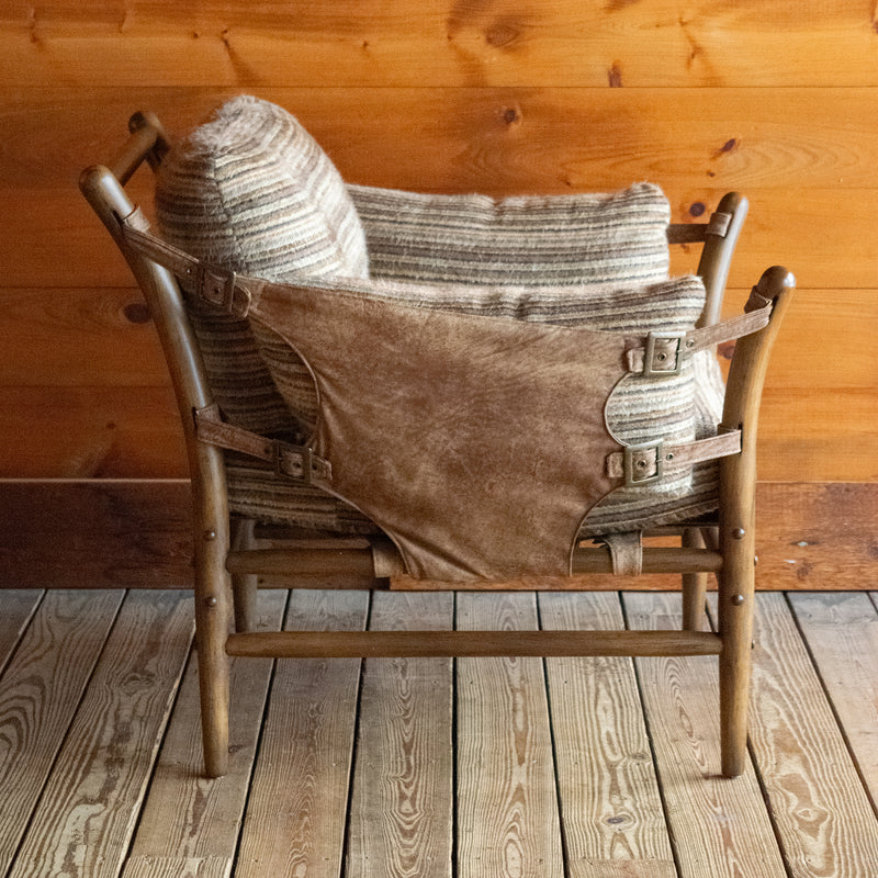 Profile view of a safari-inspired chair and ottoman with leather sides and striped cushions on a wooden floor against a wooden wall