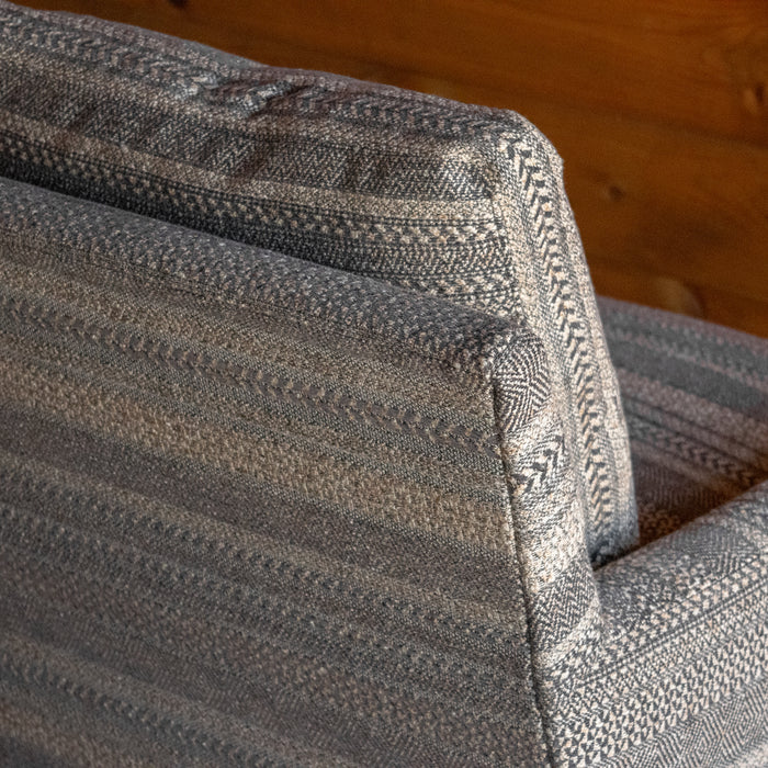 Close-up of the back of a rustic arm chair upholstered in a striped charcoal and brown fabric against a wooden wall 