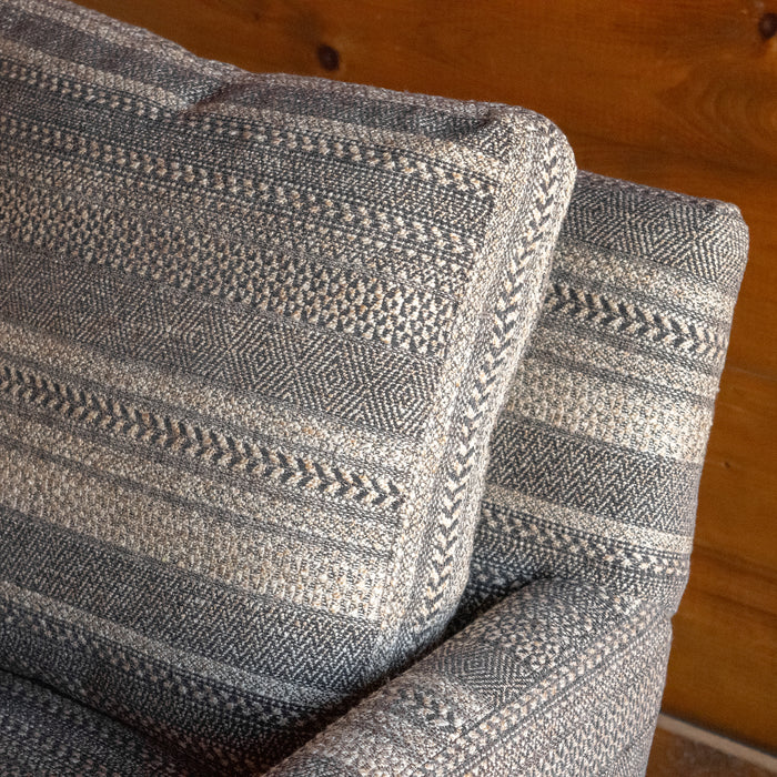 Close-up of the back cushion of a rustic arm chair upholstered in a striped charcoal and brown fabric against a wooden wall 