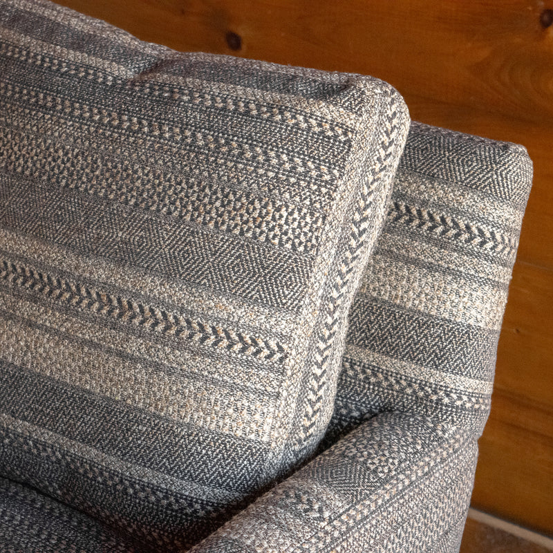 Close-up of the back cushion of a rustic arm chair upholstered in a striped charcoal and brown fabric against a wooden wall 