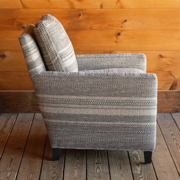 Profile view of a rustic arm chair upholstered in a striped charcoal and brown fabric with track arms and tapered wooden legs on a wooden floor against a wooden wall 