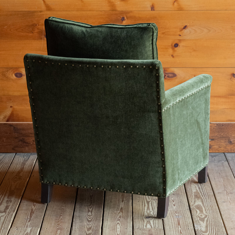 Angled back view of a rustic chair with track arms upholstered in a green velvet-chenille fabric on a wooden floor against a wooden wall