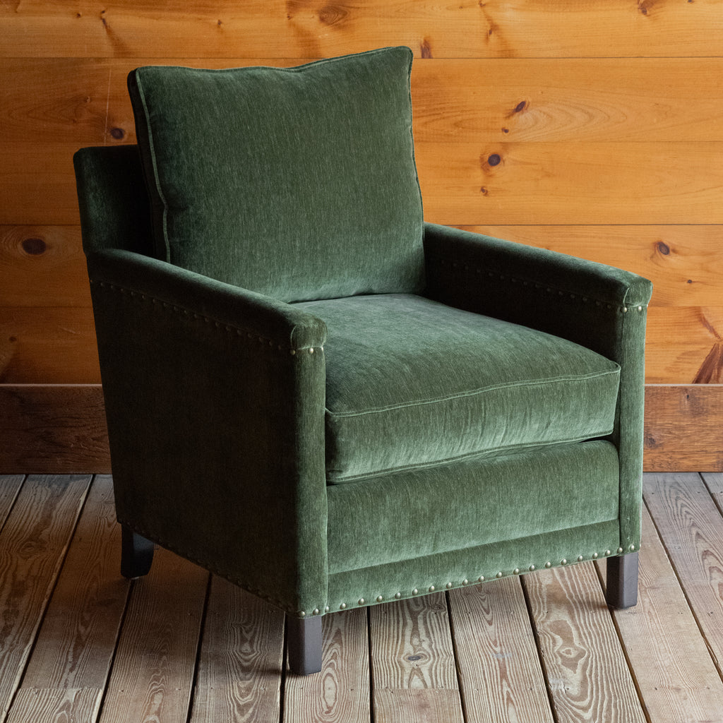 Angled front view of a rustic chair with track arms upholstered in a green velvet-chenille fabric on a wooden floor against a wooden wall