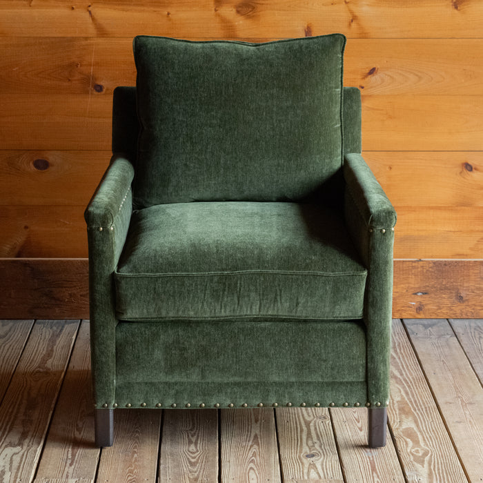 Front view of a rustic chair with track arms upholstered in a green velvet-chenille fabric on a wooden floor against a wooden wall