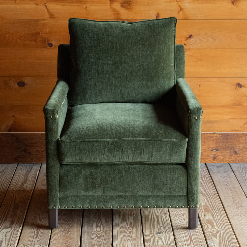 Front view of a rustic chair with track arms upholstered in a green velvet-chenille fabric on a wooden floor against a wooden wall