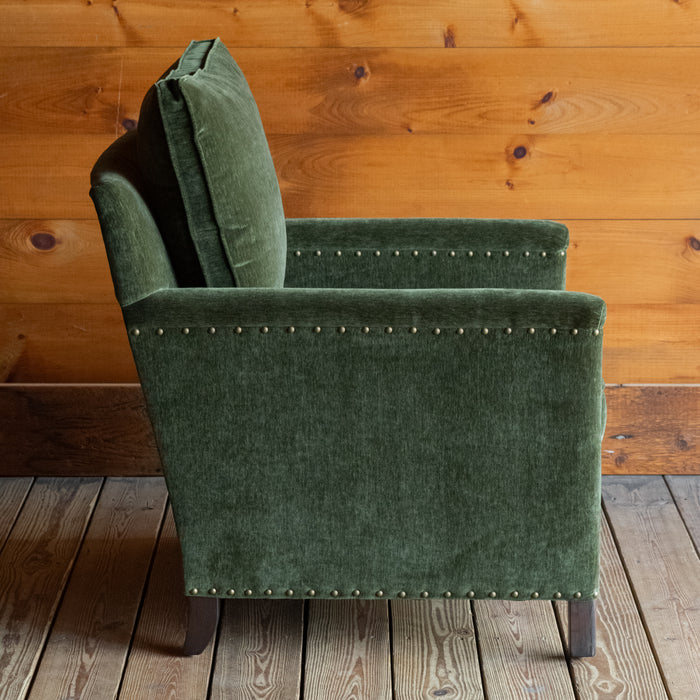 Profile view of a rustic chair with track arms upholstered in a green velvet-chenille fabric on a wooden floor against a wooden wall