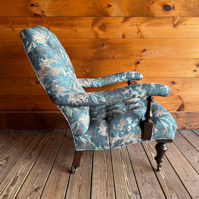 Profile view of a Victorian-inspired, open arm lounge chair on turned legs against a wooden wall.