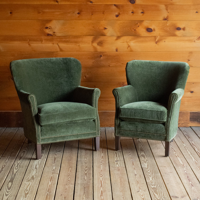 Angled back view of a large rustic arm chair upholstered in green velvet fabric on a wide-plank wooden floor against a rustic wooden wall