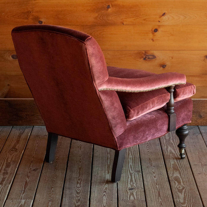 Angled back view of a warm red velvet open arm lounge chair with nailhead trim and turned wooden front legs on casters on a wooden floor against a wooden wall