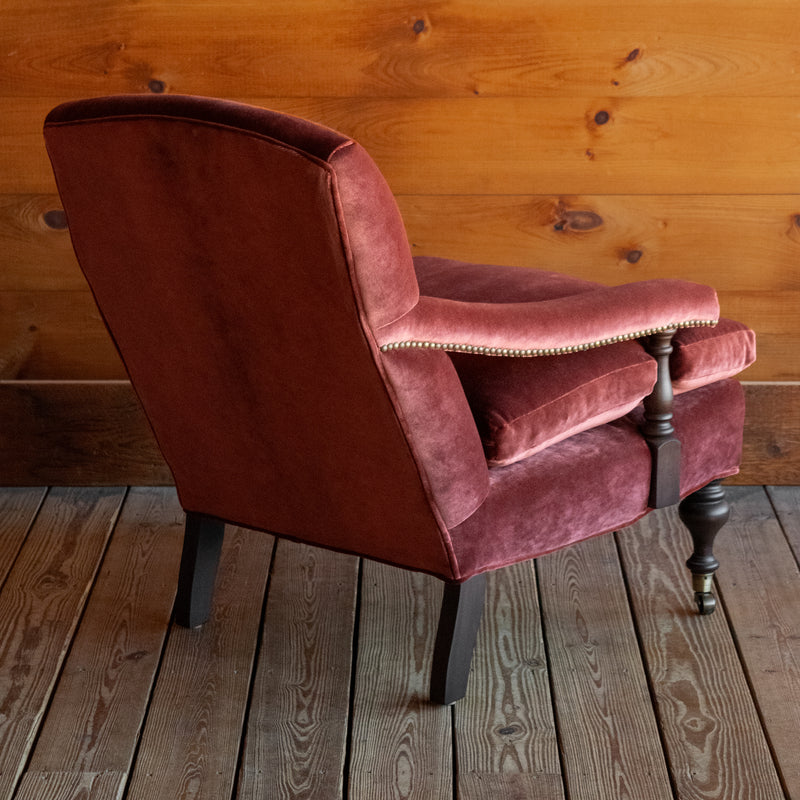 Angled back view of a warm red velvet open arm lounge chair with nailhead trim and turned wooden front legs on casters on a wooden floor against a wooden wall