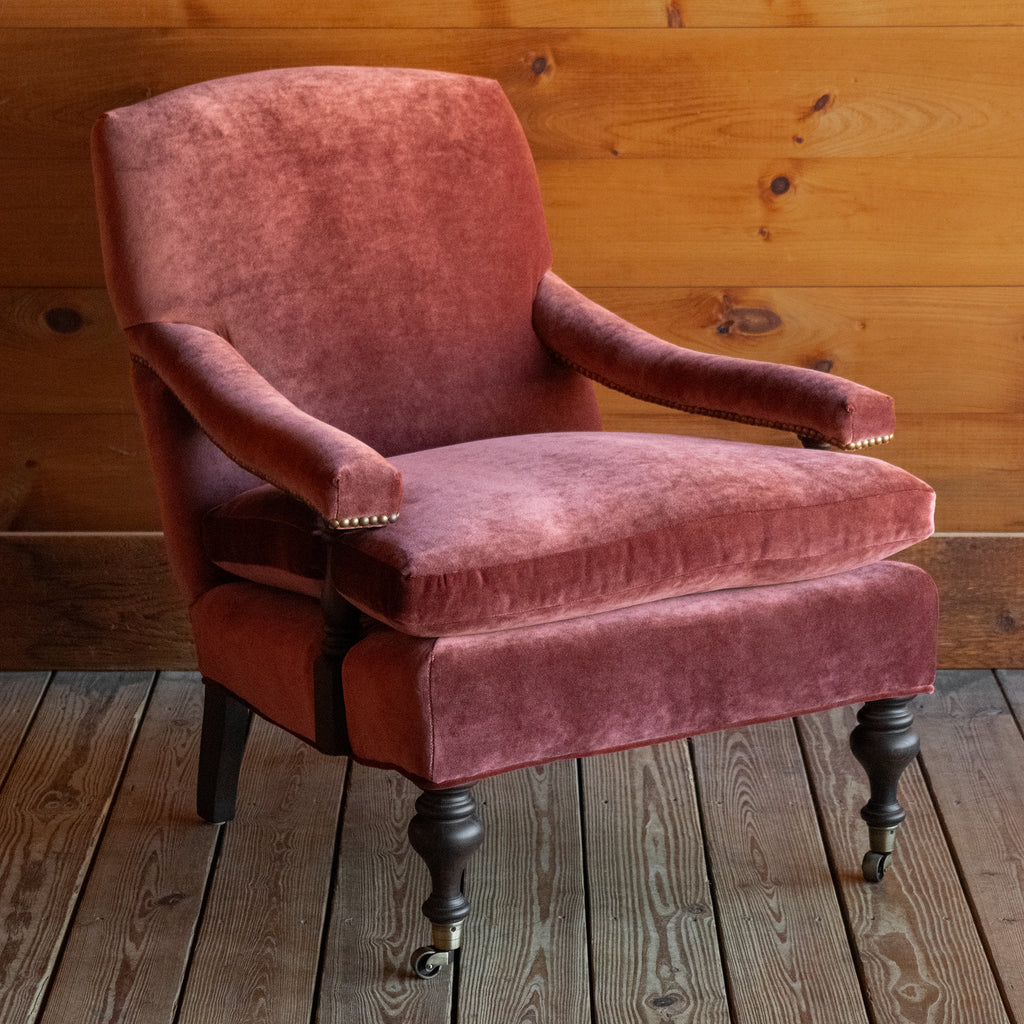 Angled front view of a warm red velvet open arm lounge chair with nailhead trim and turned wooden front legs on casters on a wooden floor against a wooden wall