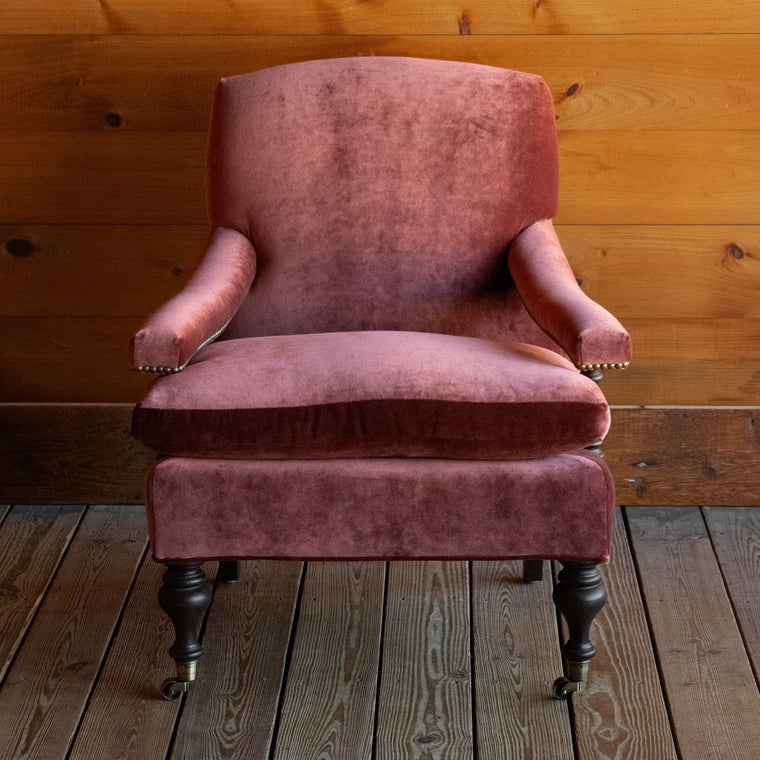 Angled back view of a warm red velvet open arm lounge chair with nailhead trim and turned wooden front legs on casters on a wooden floor against a wooden wall