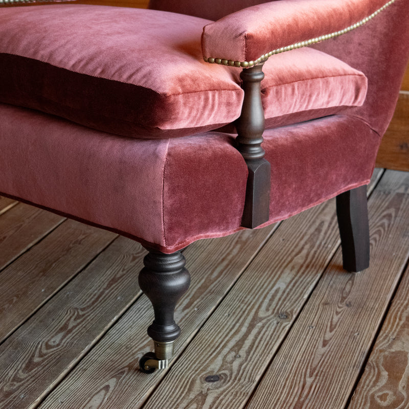 Close-up of the arm and leg of a warm red velvet open arm lounge chair with nailhead trim on a wooden floor