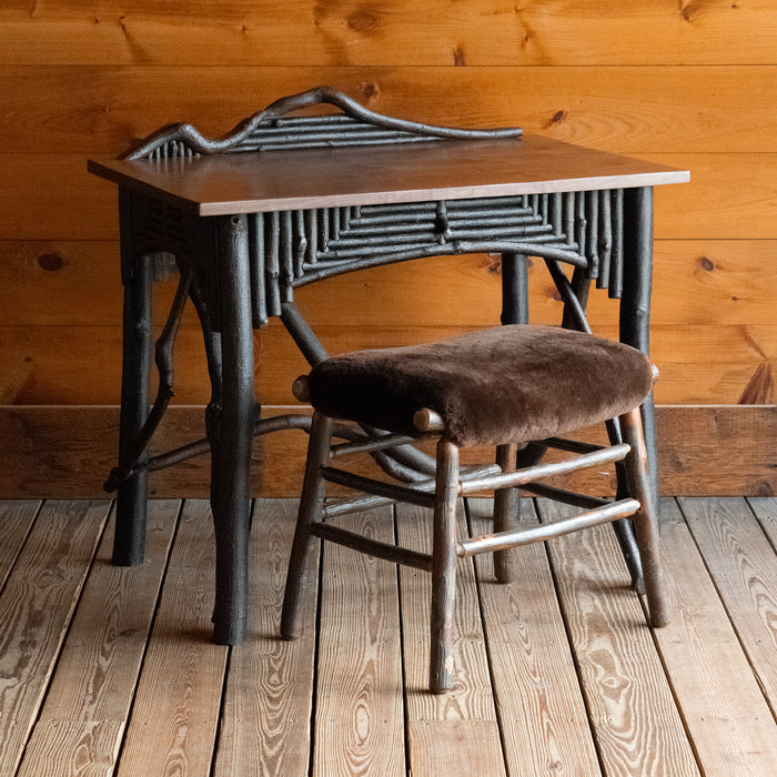Angled front view of a rustic Adirondack style desk with freeform branches and a sheepskin stool against a wooden wall 