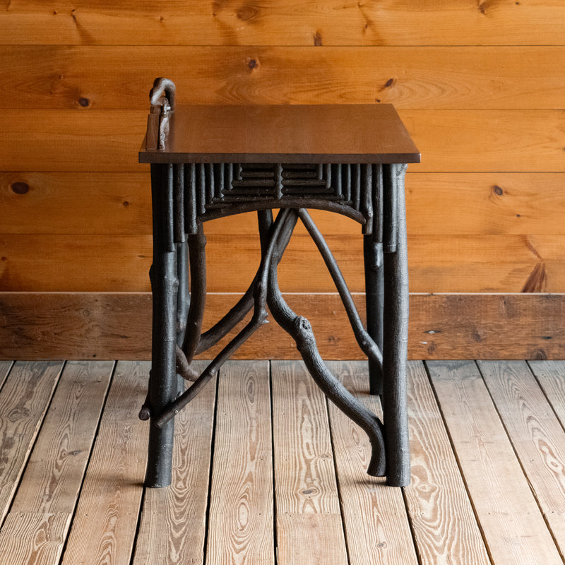 Profile view of a rustic Adirondack style desk with freeform branches and walnut top against a wooden wall 