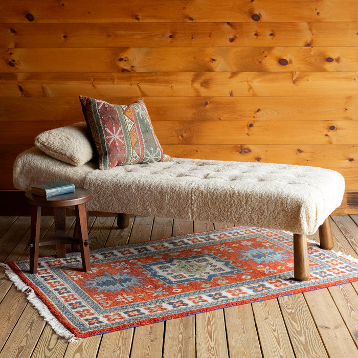 Angled front view of a chaise and matching pillow upholstered in genuine shearling with a kilim throw pillow and side table on a colorful rug atop a wide-plank wooden floor against a rustic wooden wall