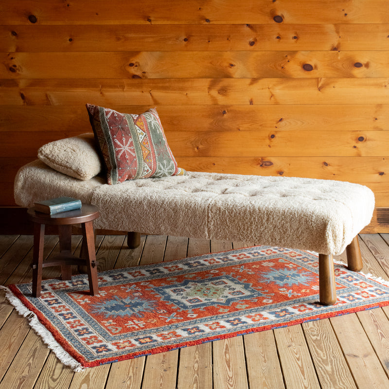 Angled front view of a chaise and matching pillow upholstered in genuine shearling with a kilim throw pillow and side table on a colorful rug atop a wide-plank wooden floor against a rustic wooden wall