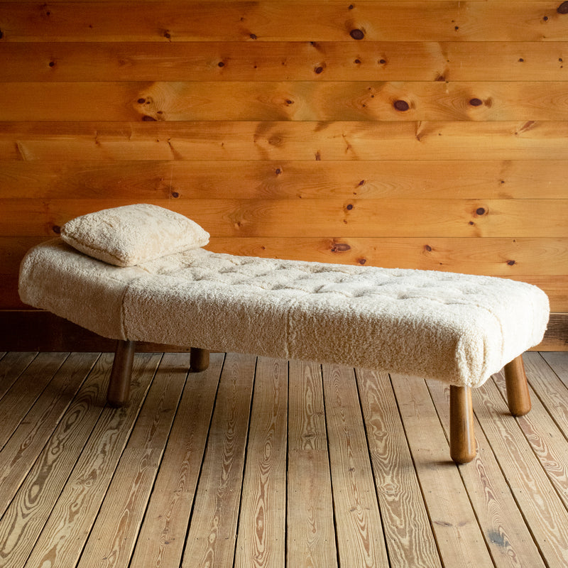 Angled front view of a chaise and matching pillow upholstered in genuine shearling on a wide-plank wooden floor against a rustic wooden wall
