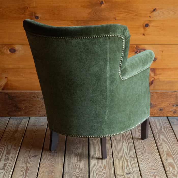 Angled back view of a petite rustic arm chair upholstered in green velvet fabric on a wide-plank wooden floor against a rustic wooden wall