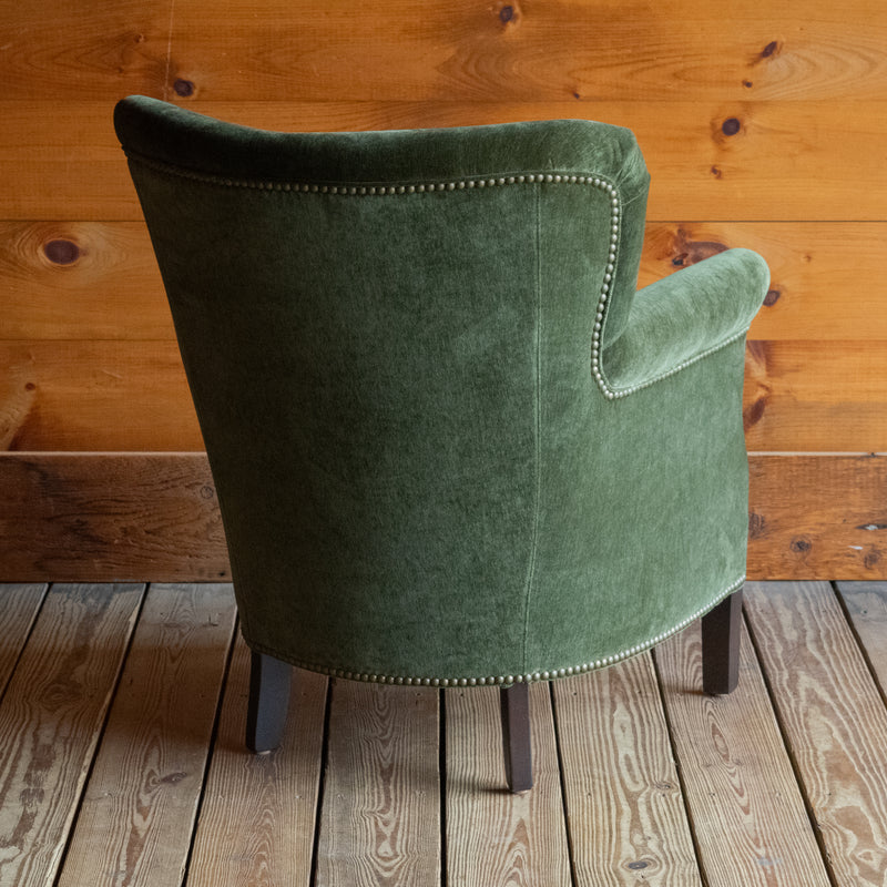 Angled back view of a petite rustic arm chair upholstered in green velvet fabric on a wide-plank wooden floor against a rustic wooden wall