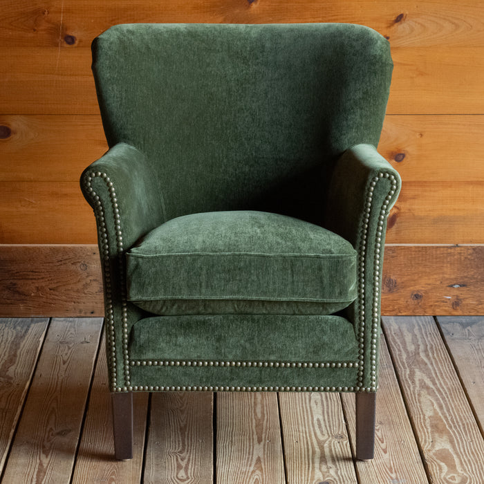 Front view of a petite rustic arm chair upholstered in green velvet fabric on a wide-plank wooden floor against a rustic wooden wall