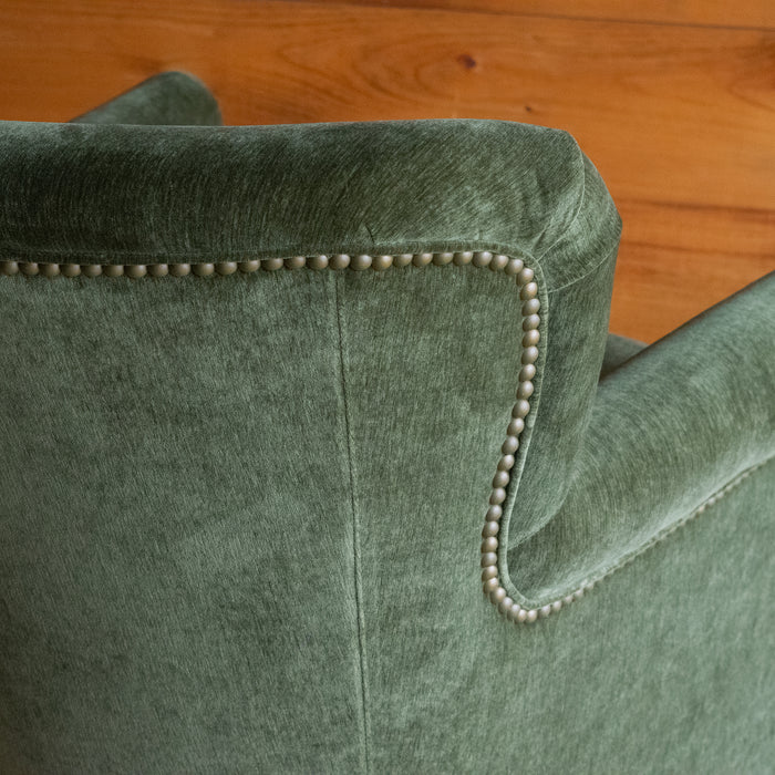Close-up of the back of a petite rustic arm chair upholstered in green velvet fabric against a rustic wooden wall