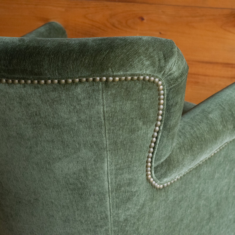 Close-up of the back of a petite rustic arm chair upholstered in green velvet fabric against a rustic wooden wall