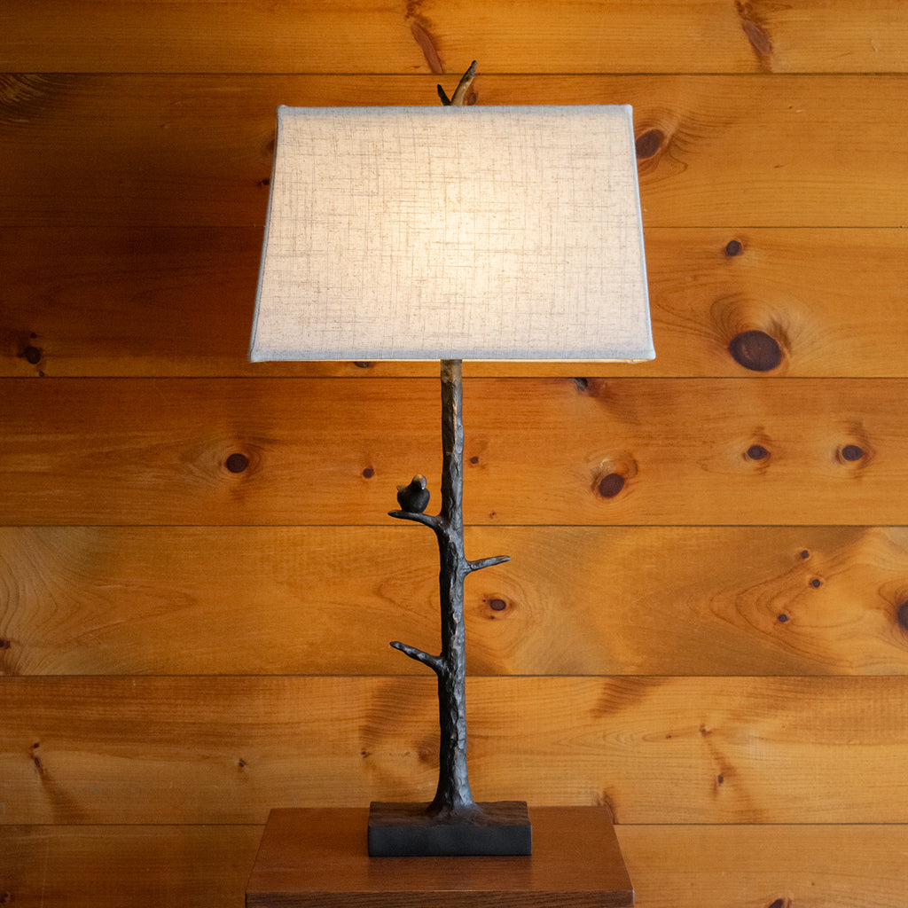 Front view of a rustic resin table lamp featuring a tree and perched bird base with a bronze metallic finish and taupe cotton shade on a wooden side table against a rustic wooden wall